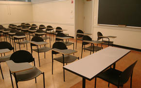 A photo of a classroom with rows of individual chairs, a table for the instructor, and a chalkboard on the side wall.