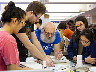 Four students and one professor leaning over a table pointing and looking at items and papers on the table. Four students and one professor leaning over a table pointing and looking at items and papers on the table.