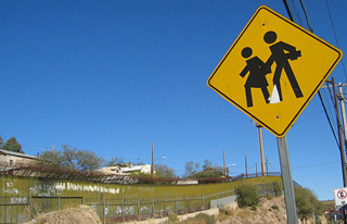 Yellow road sign featuring two figures walking together. Background is a border fence and blue sky.